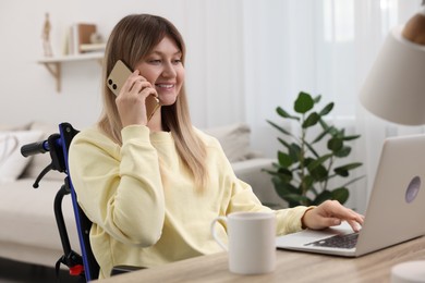 Woman in wheelchair talking on smartphone while using laptop at table in home office