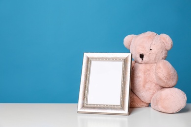 Photo of Photo frame and adorable teddy bear on table against color background, space for text. Child room elements