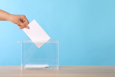 Photo of Woman putting her vote into ballot box on wooden table against light blue background, closeup. Space for text