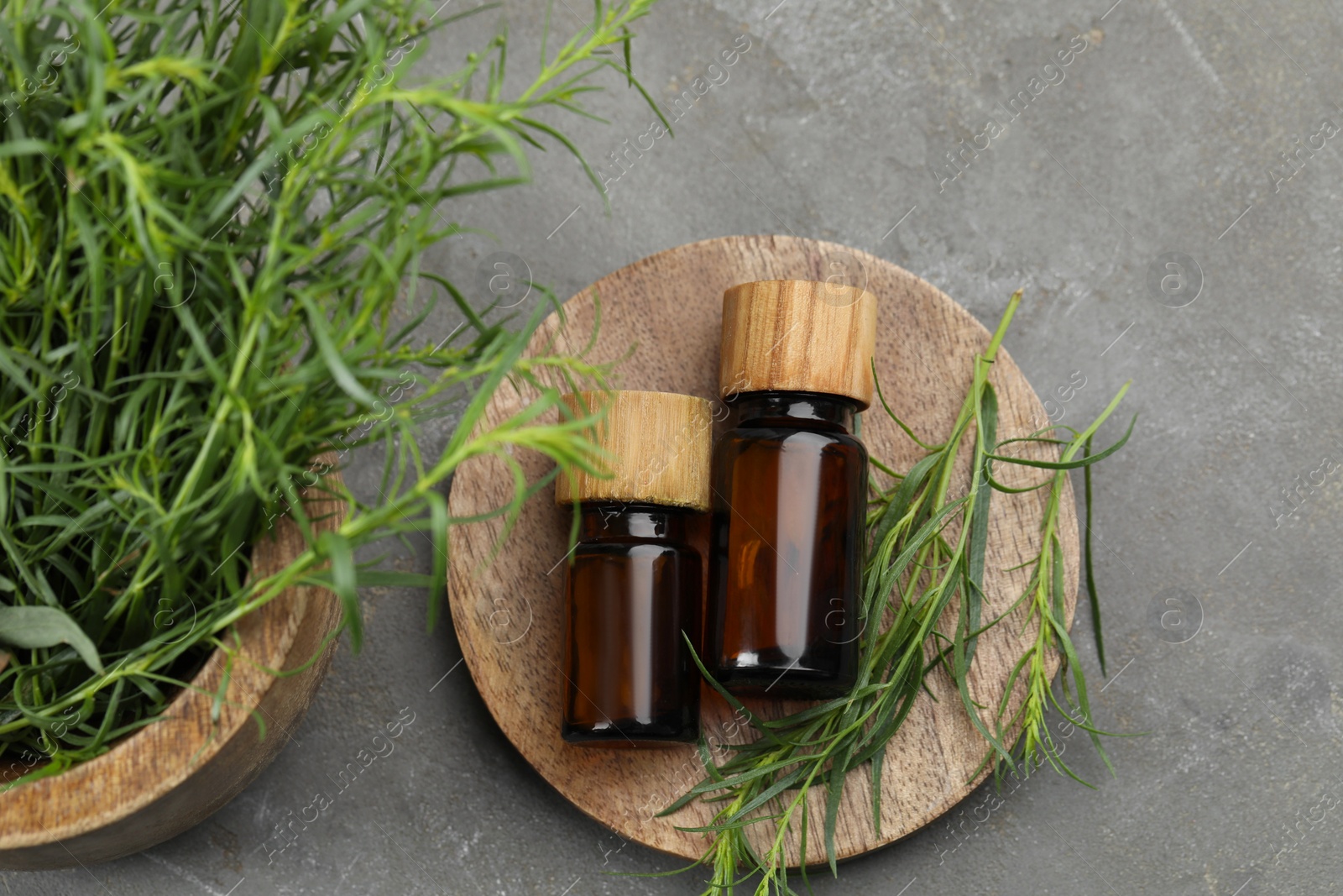 Photo of Bottles of essential oil and fresh tarragon leaves on grey table, flat lay