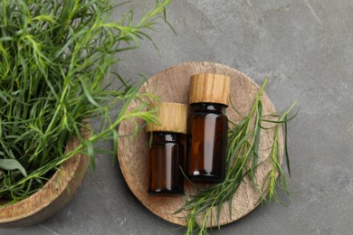 Bottles of essential oil and fresh tarragon leaves on grey table, flat lay