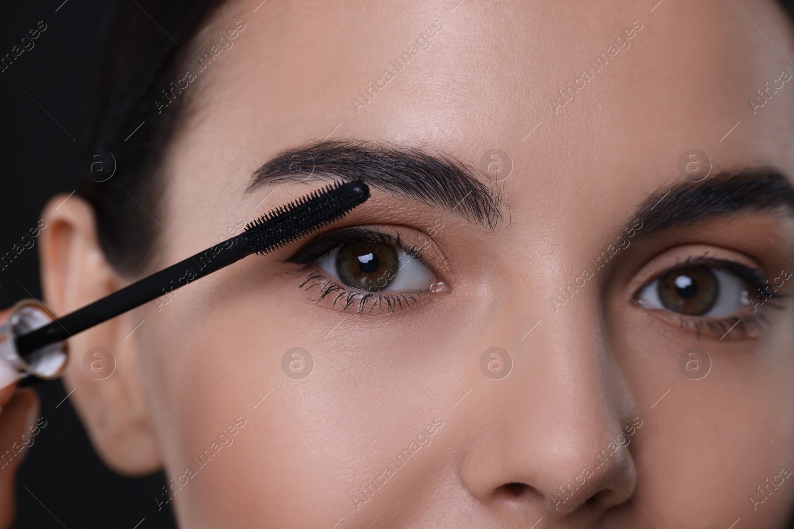 Photo of Beautiful young woman applying mascara on black background, closeup