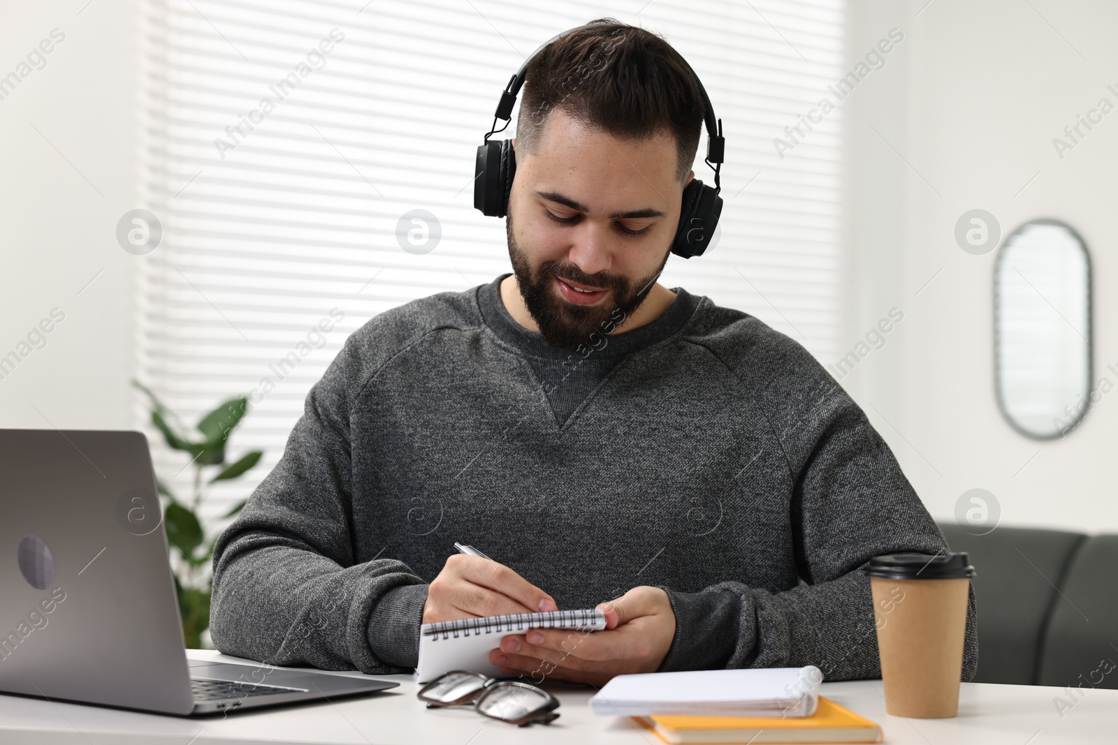 Photo of E-learning. Young man taking notes during online lesson at white table indoors