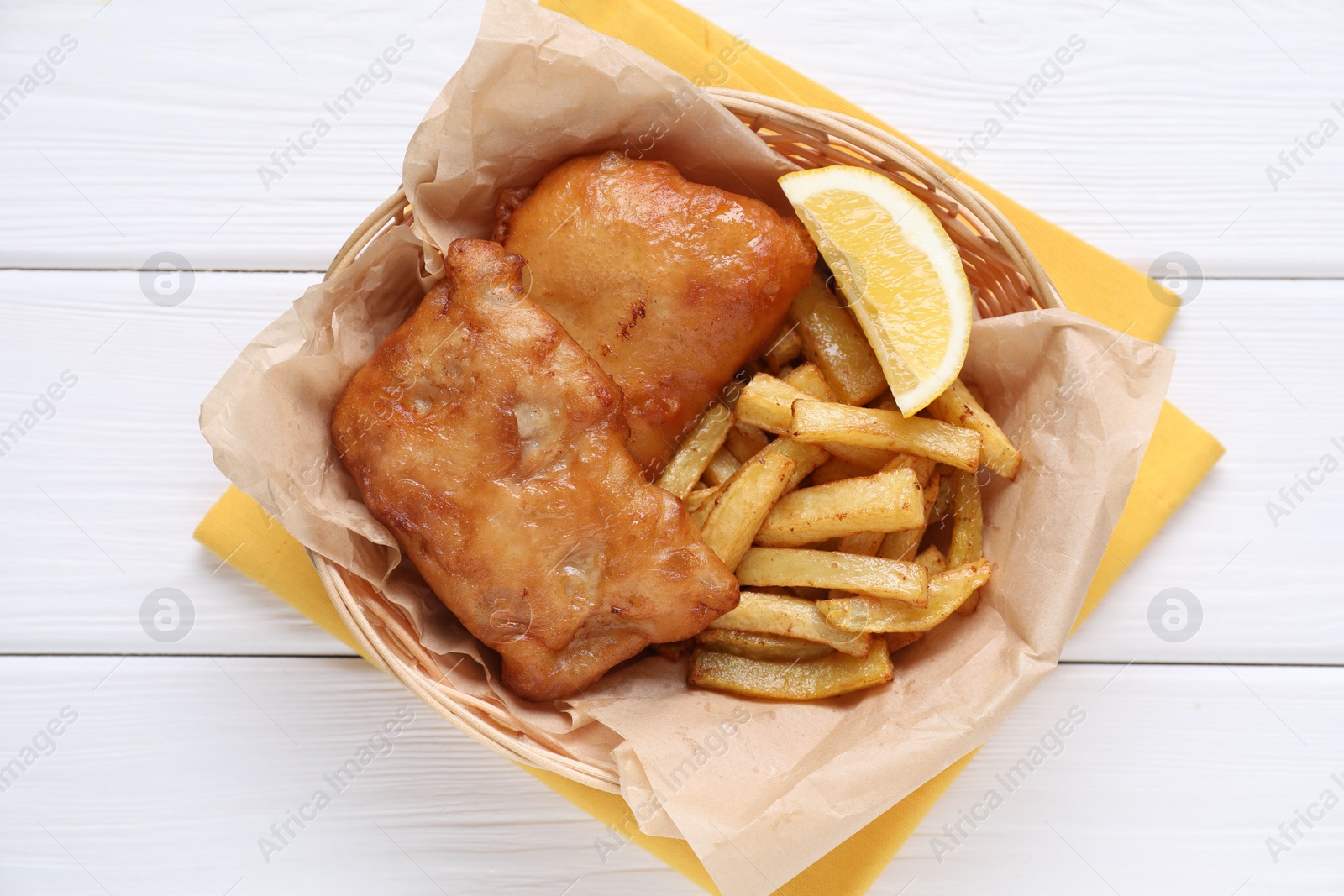 Photo of Tasty fish, chips and lemon in wicker bowl on white wooden table, top view