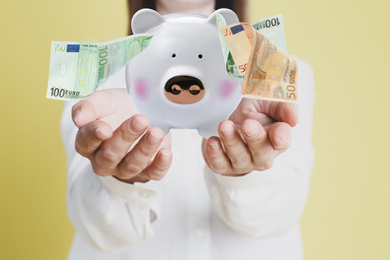 Young woman holding piggy bank and Euro banknotes falling on color background, closeup 