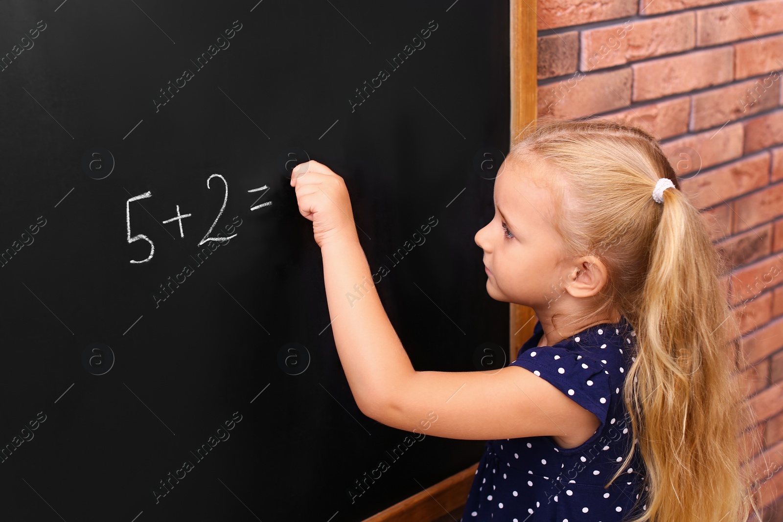 Photo of Cute little left-handed girl doing sums on chalkboard near brick wall