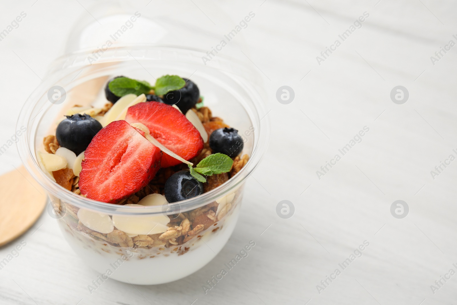 Photo of Tasty granola with berries, yogurt and almond flakes in plastic cup on white wooden table, closeup. Space for text