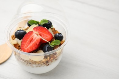 Photo of Tasty granola with berries, yogurt and almond flakes in plastic cup on white wooden table, closeup. Space for text