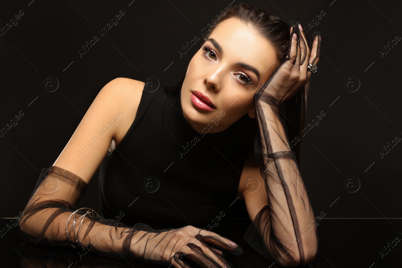 Photo of Beautiful young woman in evening gloves at black glass table against dark background