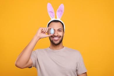 Photo of Happy African American man in bunny ears headband covering eye with Easter egg on orange background. Space for text