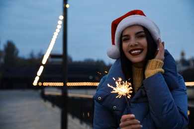 Photo of Woman in Santa hat holding burning sparkler outdoors, space for text