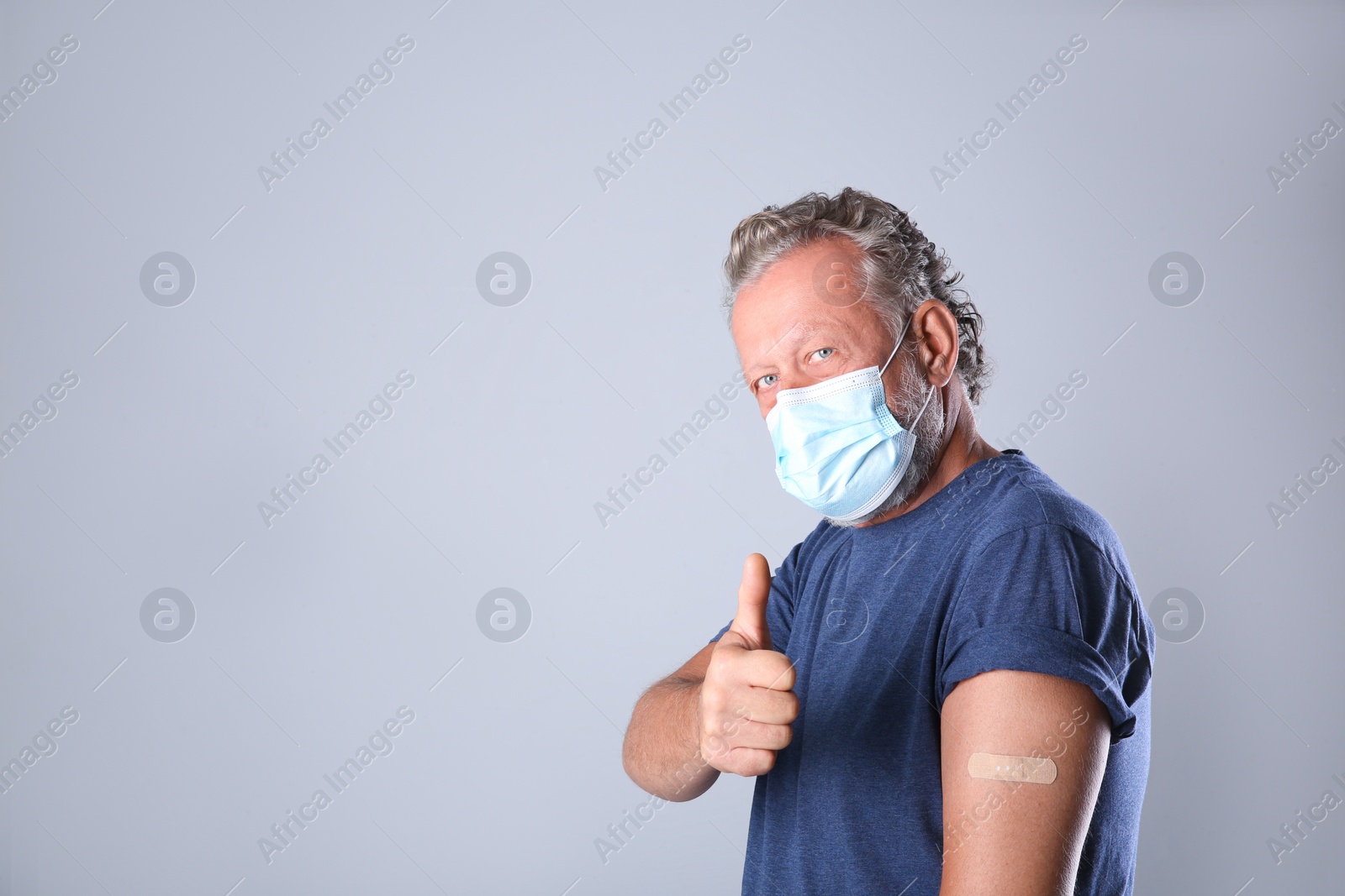 Photo of Cheerful senior man in protective mask showing arm with bandage after vaccination on grey background. Space for text
