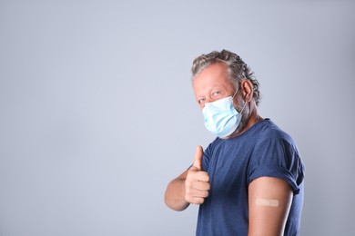 Cheerful senior man in protective mask showing arm with bandage after vaccination on grey background. Space for text