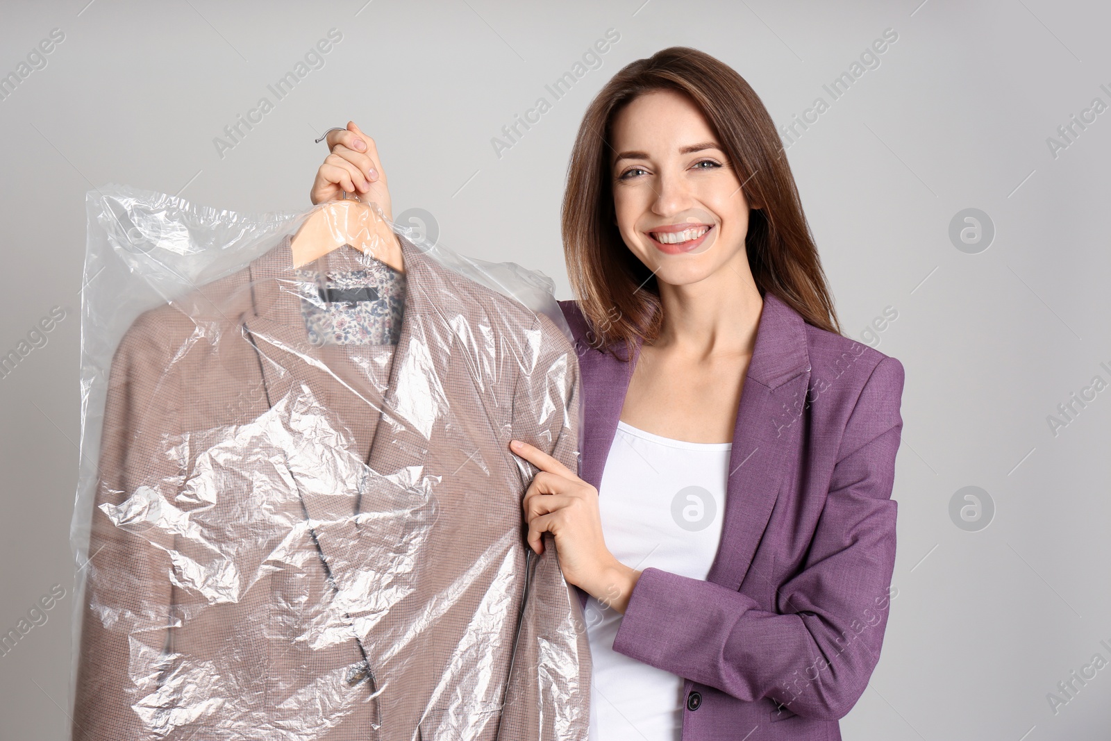 Photo of Young woman holding hanger with jacket in plastic bag on grey background. Dry-cleaning service