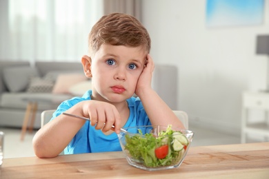 Photo of Emotional little boy eating vegetable salad at table in room