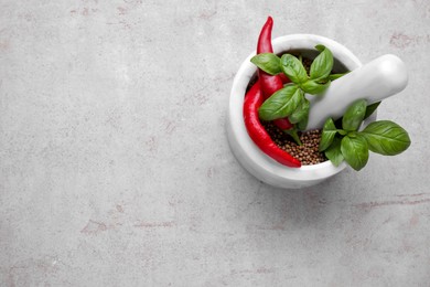 Mortar with peppercorns, basil and chilli pepper on light grey table, top view. Space for text