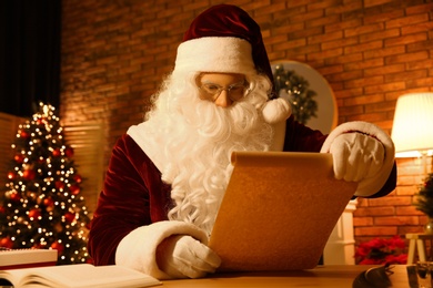 Photo of Santa Claus reading wish list at table indoors