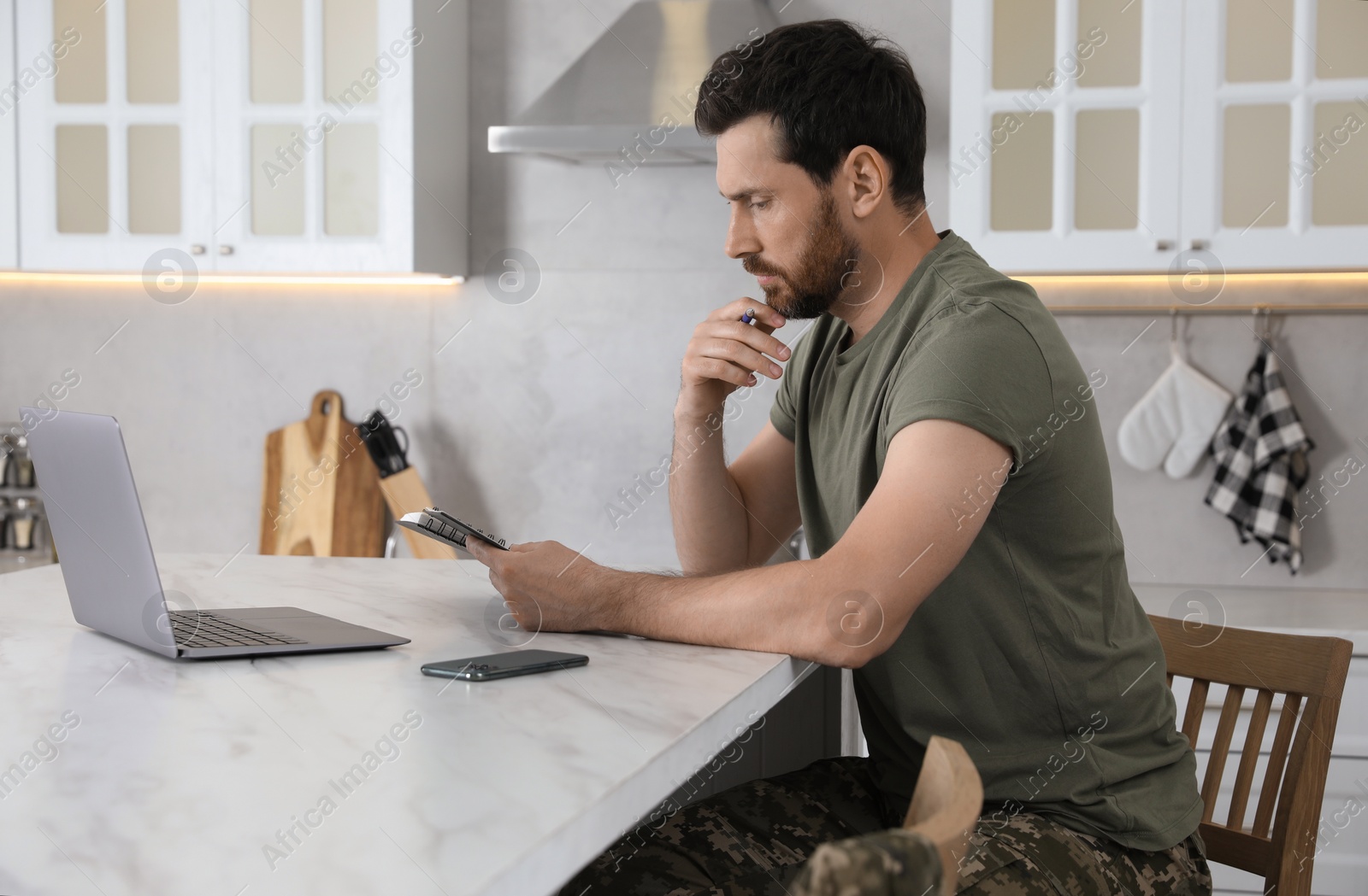 Photo of Soldier taking notes while working with laptop at white marble table in kitchen. Military service