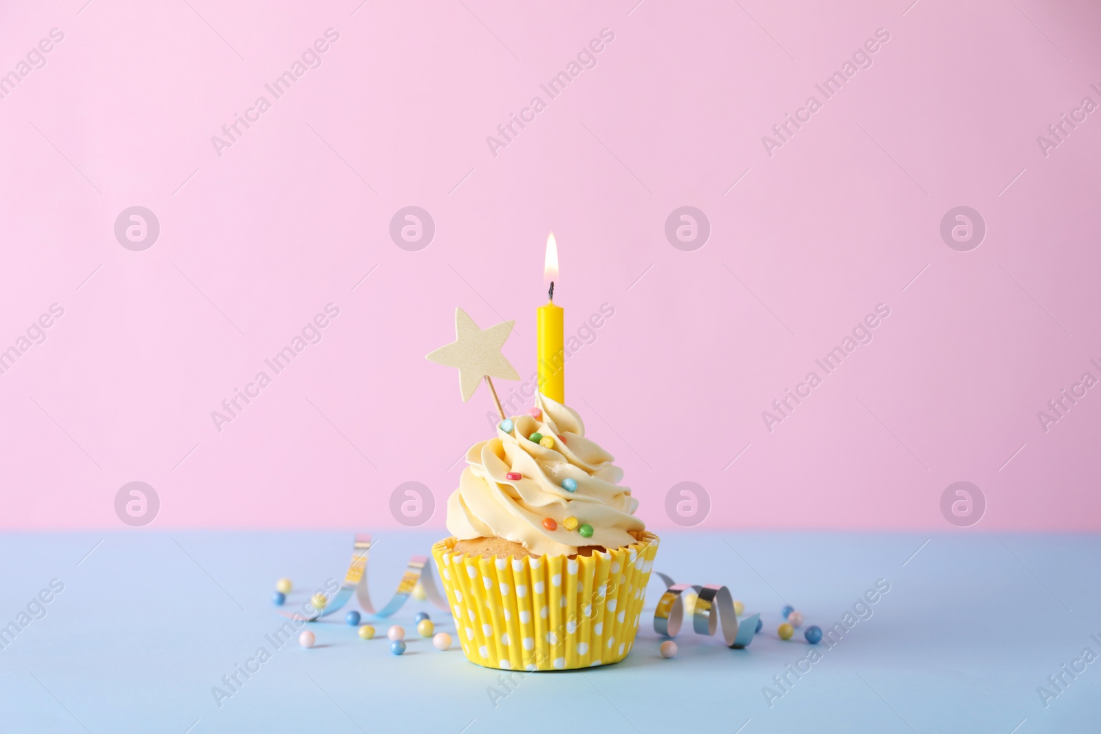 Photo of Delicious birthday cupcake with burning candle, sprinkles and streamer on light blue table against pink background