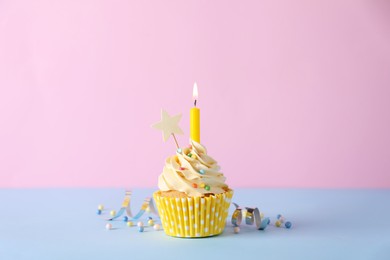 Delicious birthday cupcake with burning candle, sprinkles and streamer on light blue table against pink background