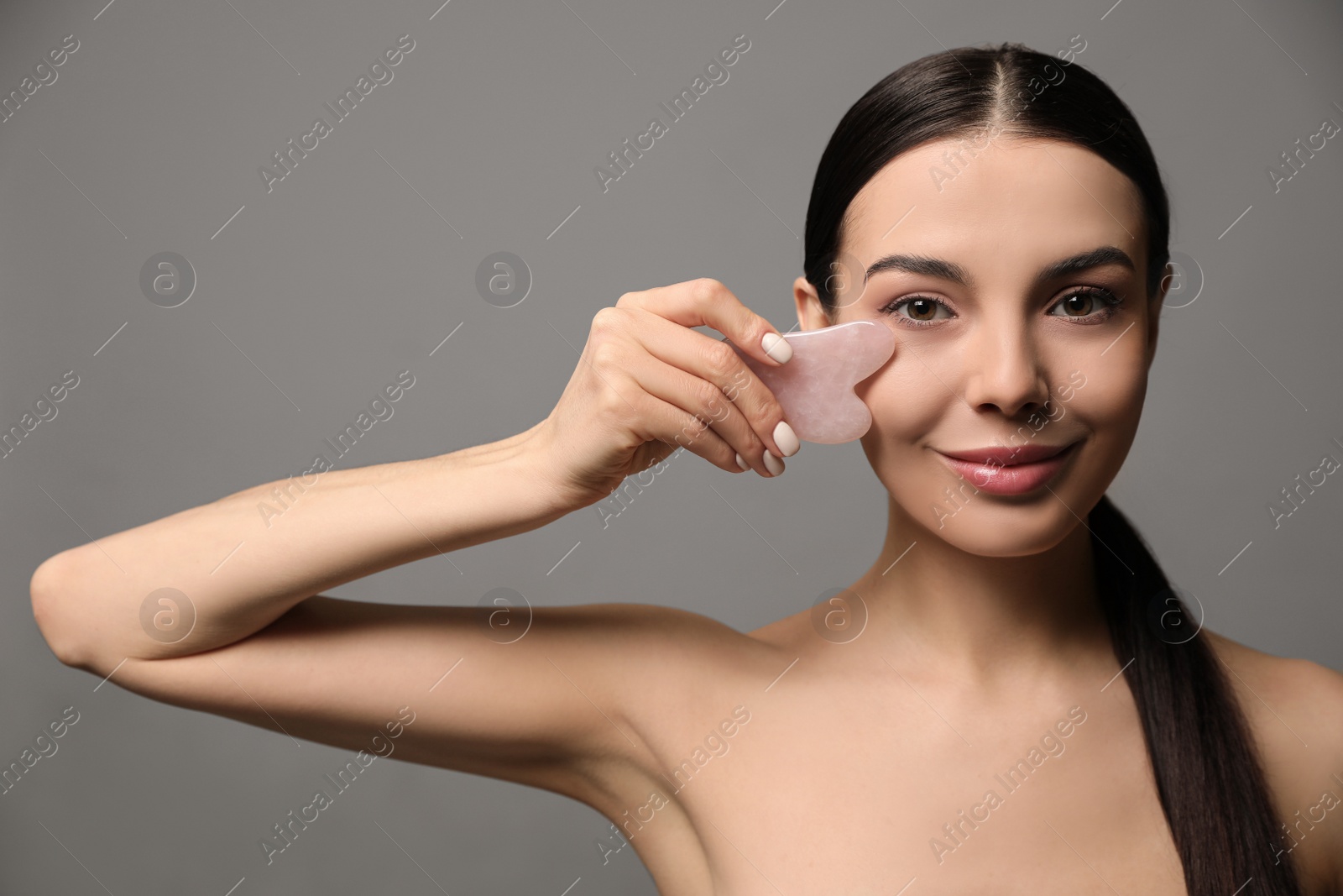 Photo of Beautiful young woman doing facial massage with gua sha tool on grey background