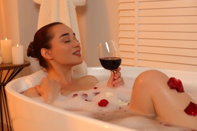 Photo of Woman with glass of wine taking bath in tub with foam and rose petals indoors