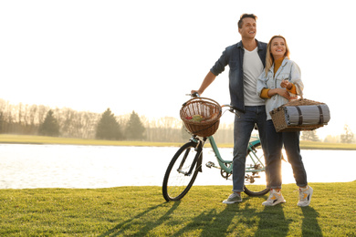 Young couple with bicycle and picnic basket near lake on sunny day