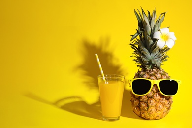 Glass of fresh pineapple juice and fruit with sunglasses on yellow background, space for text