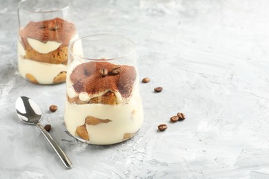 Delicious tiramisu in glasses, coffee beans and spoon on grey textured table, space for text