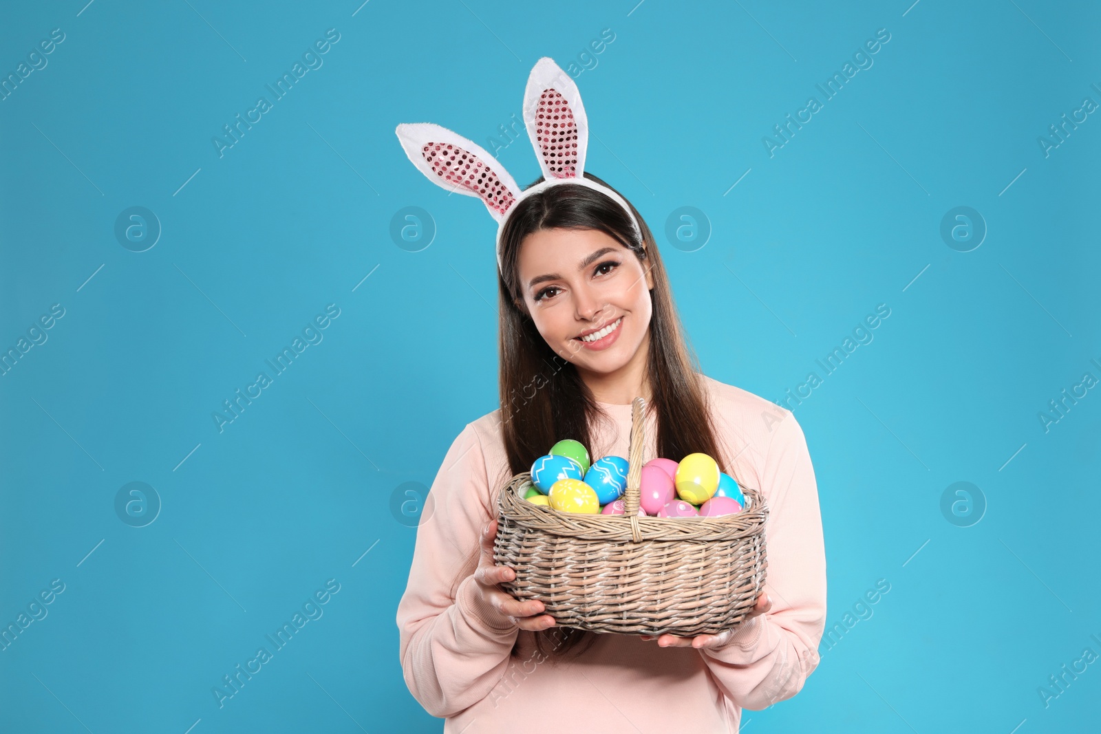 Photo of Beautiful woman in bunny ears headband holding basket with Easter eggs on color background