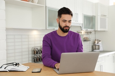 Photo of Handsome young man working with laptop at table in kitchen
