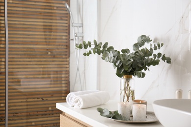 Photo of Fresh eucalyptus branches and burning candles on countertop in bathroom