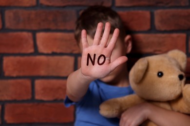 Child abuse. Boy with teddy bear making stop gesture near brick wall, selective focus. No written on his hand