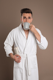 Young man in bathrobe with cup of coffee on brown background