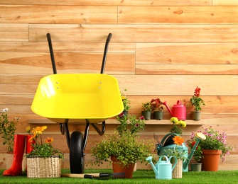 Wheelbarrow with gardening tools and flowers near wooden wall