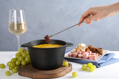 Photo of Woman dipping piece of bread into fondue pot with tasty melted cheese at white wooden table against gray background, closeup