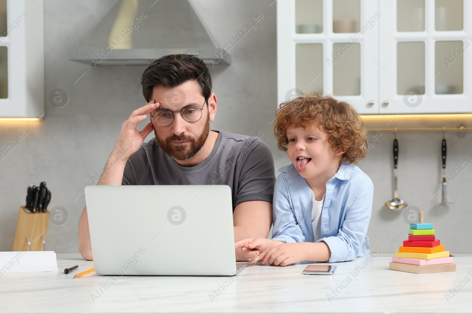 Photo of Little boy bothering father while he working remotely at home. Man with laptop and his child at desk in kitchen