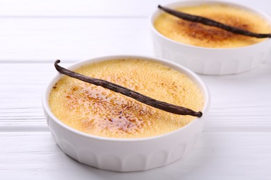 Photo of Delicious creme brulee in bowls and vanilla pods on white wooden table, closeup
