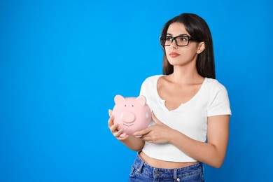 Photo of Young woman in eyeglasses with piggy bank on light blue background, space for text