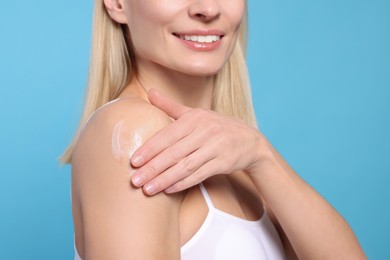 Woman applying body cream onto her arm against light blue background, closeup