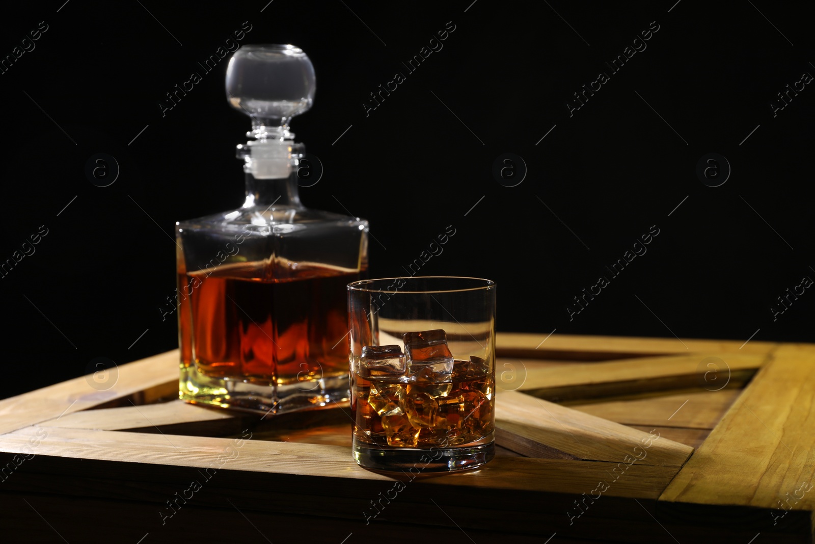 Photo of Whiskey with ice cubes in glass and bottle on wooden crate against black background, space for text