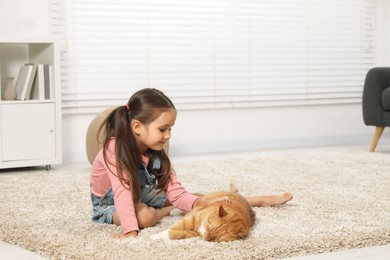 Photo of Smiling little girl petting cute ginger cat on carpet at home, space for text