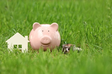 Photo of Piggy bank, house figure and jar with coins on green grass in park. Space for text