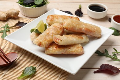 Photo of Tasty fried spring rolls, lime, sauces and other products on light wooden table, closeup