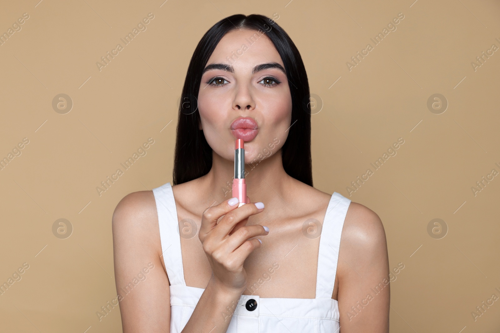 Photo of Young woman with beautiful makeup holding nude lipstick on beige background