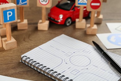 Many different road signs, notebook with sketch of roundabout and toy car on wooden table, closeup. Driving school