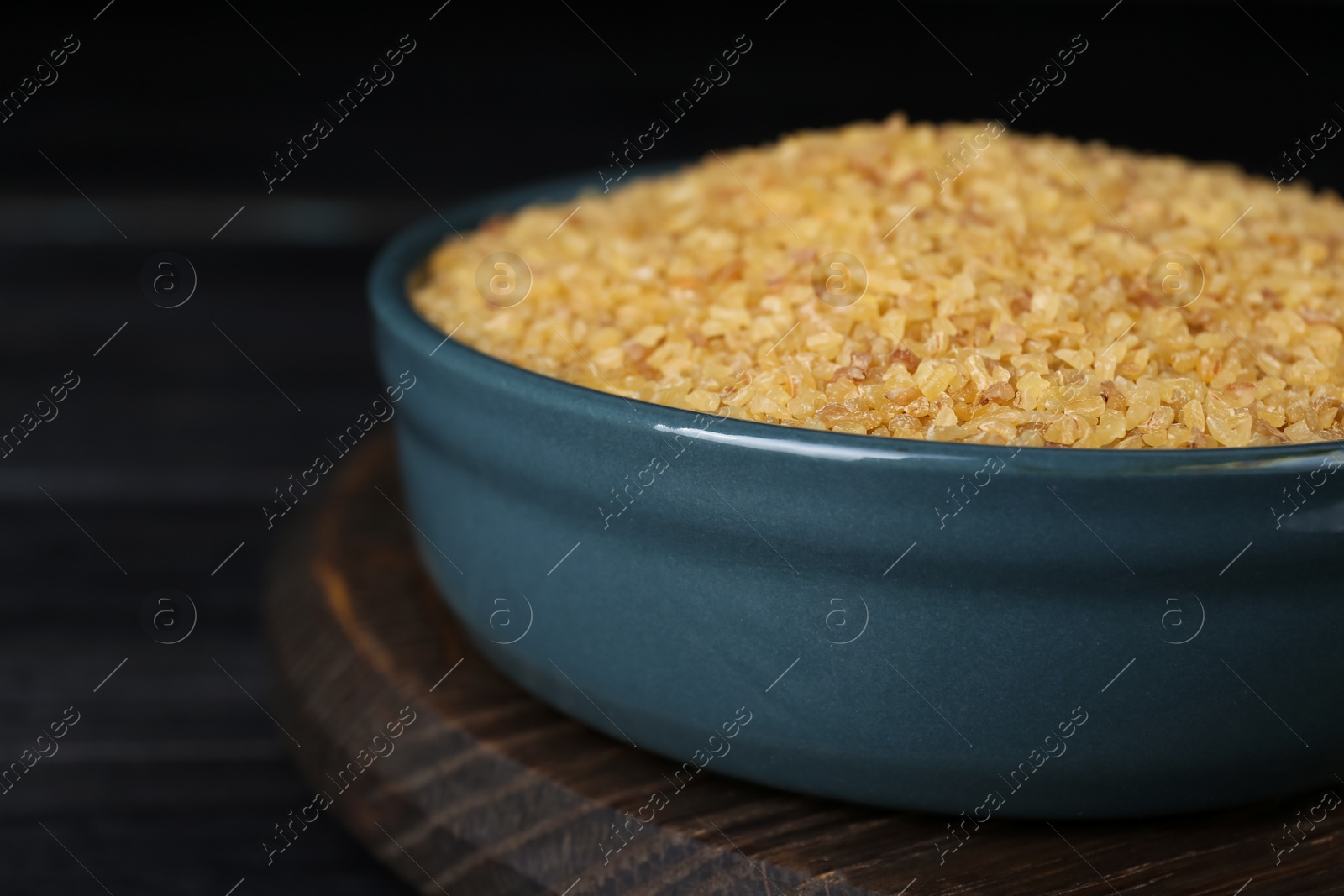 Photo of Bowl of uncooked bulgur on black wooden table, closeup