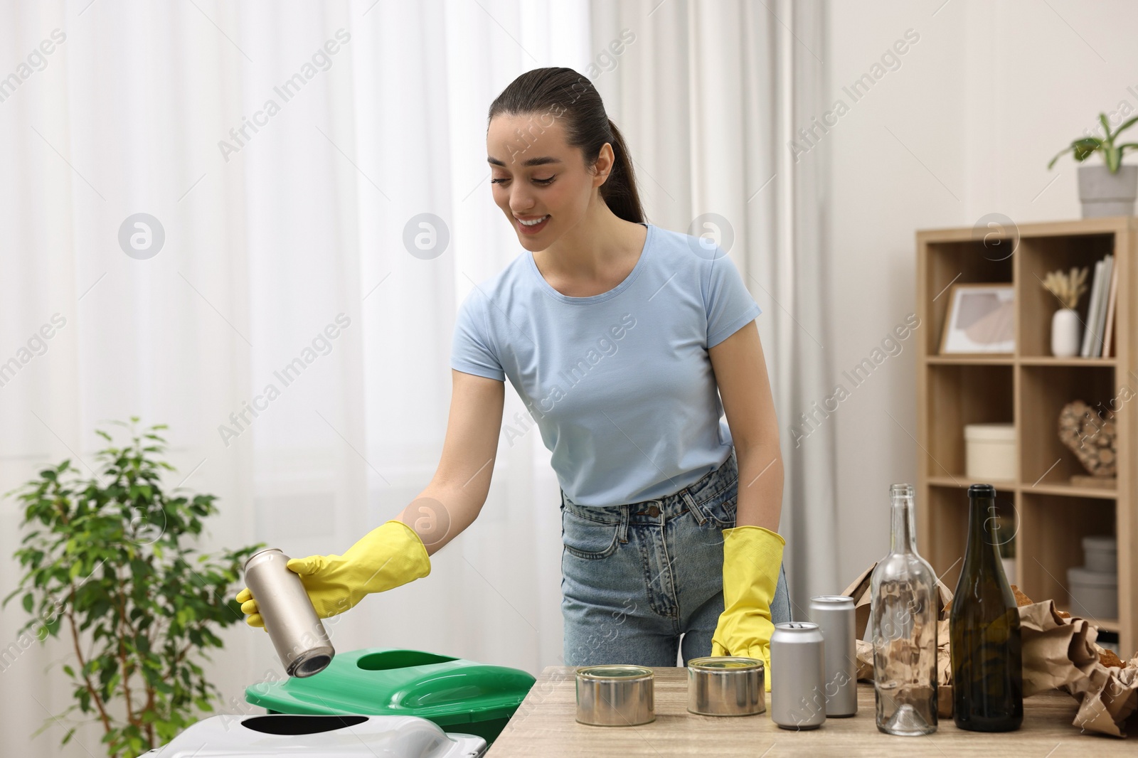 Photo of Garbage sorting. Smiling woman throwing metal can into trash bin in room