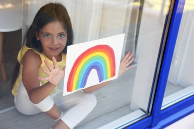 Photo of Little girl with picture of rainbow near window, view from outdoors. Stay at home concept
