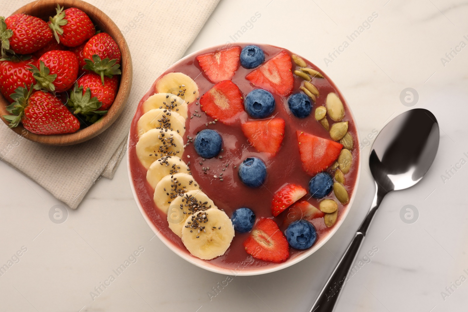 Photo of Bowl of delicious smoothie with fresh blueberries, strawberries, banana and pumpkin seeds on white table, flat lay